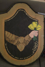 Device: Sable, a sinister arm fesswise reversed embowed proper armored Or grasping three tulips Or slipped vert all within a bordure Or.

About the additional text on my shield: I have always liked the song 'Tub-Thumping' by Chumbawamba. While it is about politicians who are 'stumping' (The term used here in America) for election I took it as a personal theme song when my health tanked a few years ago. So with a bit of help from Google and Babel translations on-line I give you:

Ego Adepto infligo d